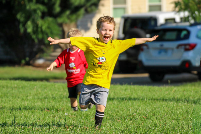A boy in a yellow jersey cheering with arms spread wide at a HappyFeet soccer class