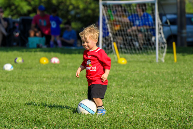 Toddler in a red jersey dribbling a soccer ball at a HappyFeet class near Overland Park, KS