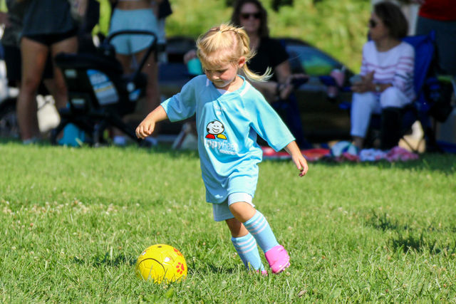 Toddler girl in a light blue jersey approaching a yellow soccer ball at a HappyFeet class in Parkville, MO