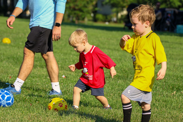 Two toddler boys in red and yellow competing for the ball as a coach guides them at a HappyFeet class in Leawood, KS